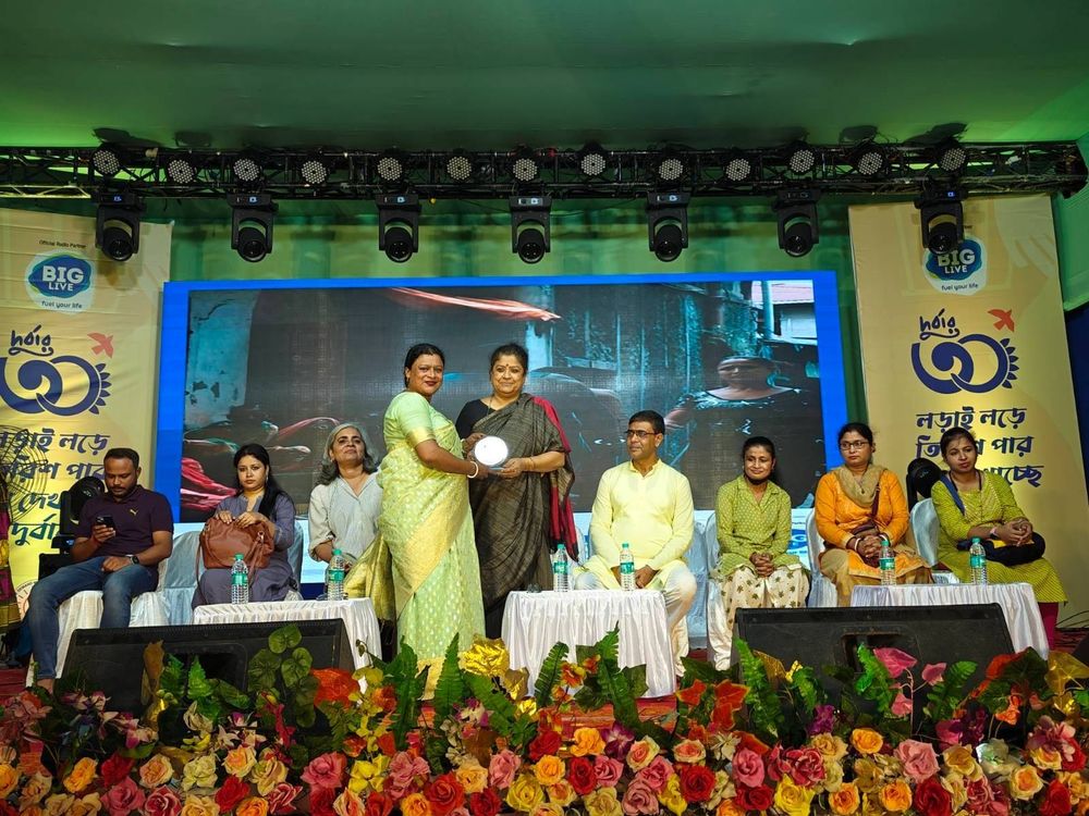 Nine people seated on a flower‑lined stage at the Durbar Mahila Samanwaya Committee (DMSC) 30th anniversary. At center, a DMSC representative presents an award to CREA’s Executive Director (India) while others sit to either side. Event banners and a large screen form the backdrop