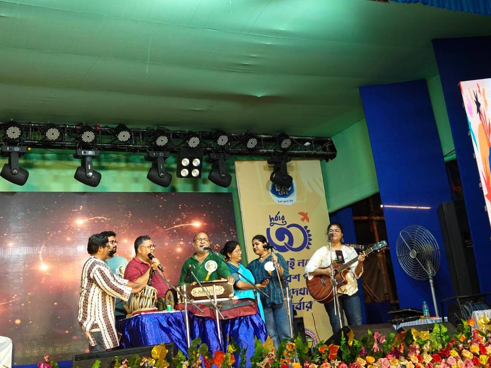 A group of musicians performing live on stage during the DMSC 30th anniversary celebration. They are playing instruments like the tabla and guitar, with singers using microphones. The stage is adorned with bright floral decorations and a digital backdrop