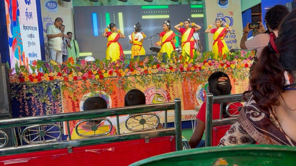 A group of young girls in yellow and red traditional attire performing a cultural dance on stage, decorated with vibrant flowers, during the DMSC 30th anniversary celebration. Audience members watch from the front rows while a person records the performance on a phone