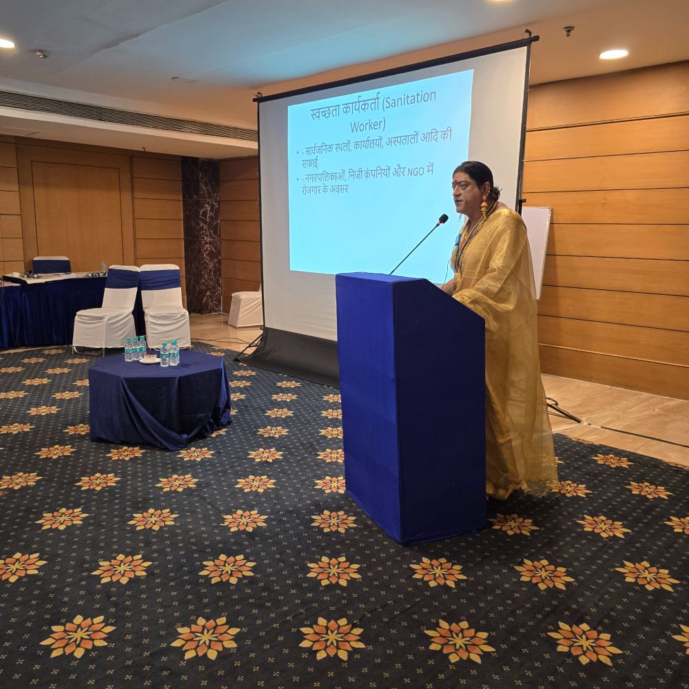 Image of a speaker at the lectern with a white screen behind them. The speaker is wearing a golden saree and is standing on a carpeted floor