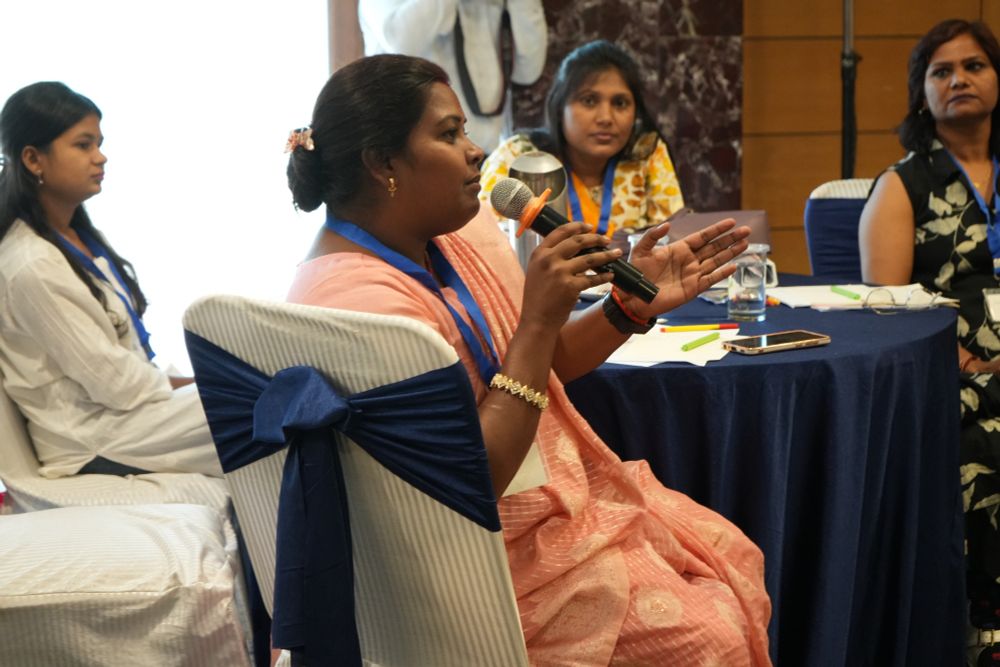 Participants sitting around a table, the one in the middle is wearing a pink saree and speaking into the mic, the other three are attentively listening.