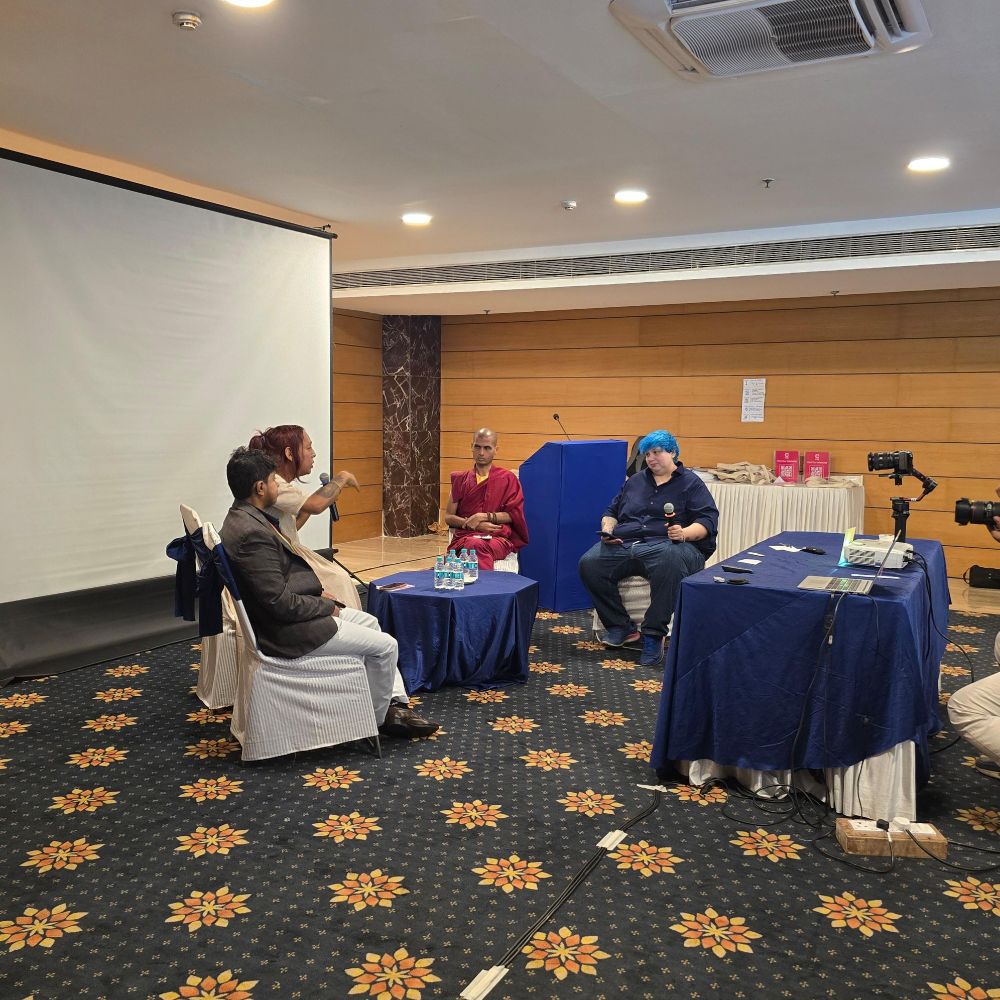 
Image of four panelists sitting in a semicircle on white chairs. They have a round blue table between them. There table on the side of the frame with a camera mounted on it and there's an empty white screen behind the panelists