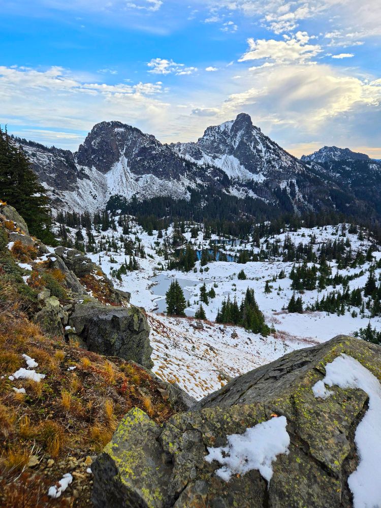 The mountains of the Central Cascades of Washington state,  lightly dusted in snow in mid-October, with autumn colors peeking through in places that receive plenty of sun. There's a crystal blue alpine lake that isn't frozen over quite yet and several mountain tarns that are frozen over with a thin dusting of snow over the ice. The sky is blue with some light cumulus and cirrus clouds, for the perfect mountain scene. 