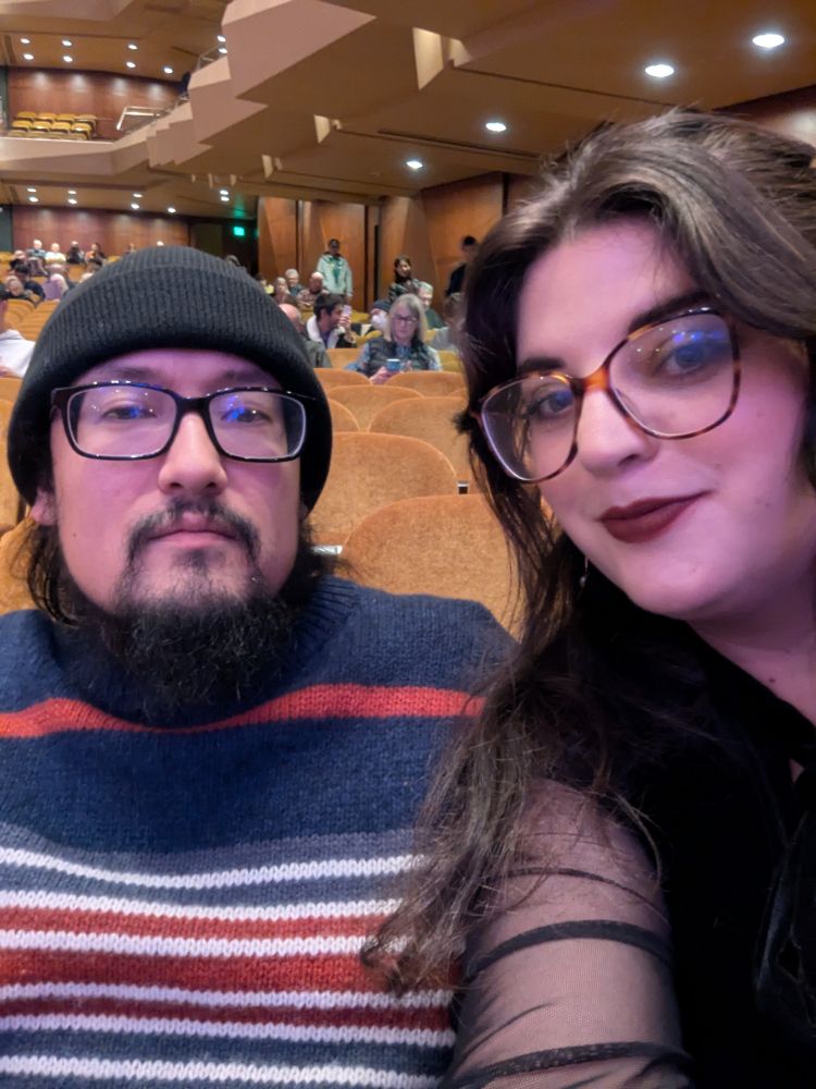 A man and woman in glasses and date night outfits sitting in the audience before the symphony. 