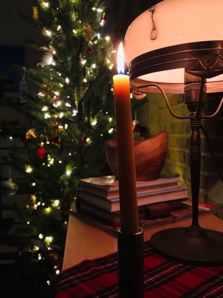 A christmas scene: a lit taper candle in the foreground in front of a white dome lamp and stack of books with a christmas tree with white lights in thr background