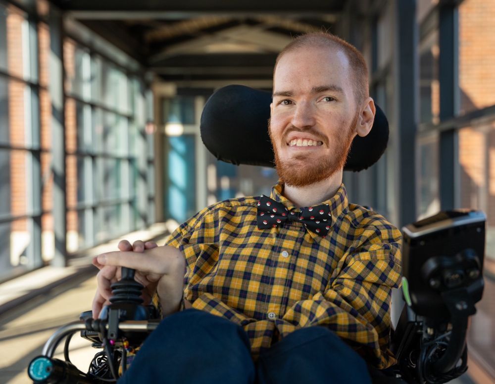 Grant smiling at the camera. He is wearing a checkered button shirt, a Pokéball bow tie, and blue dress pants. He is sitting in an electric wheelchair.