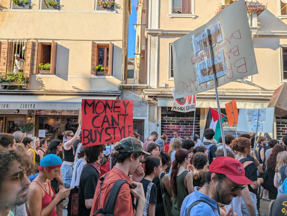 Protest march in a high street in Venice, Italy. A crowd of good looking, well dressed young people, some carrying placards - one is turned towards the camera reading 