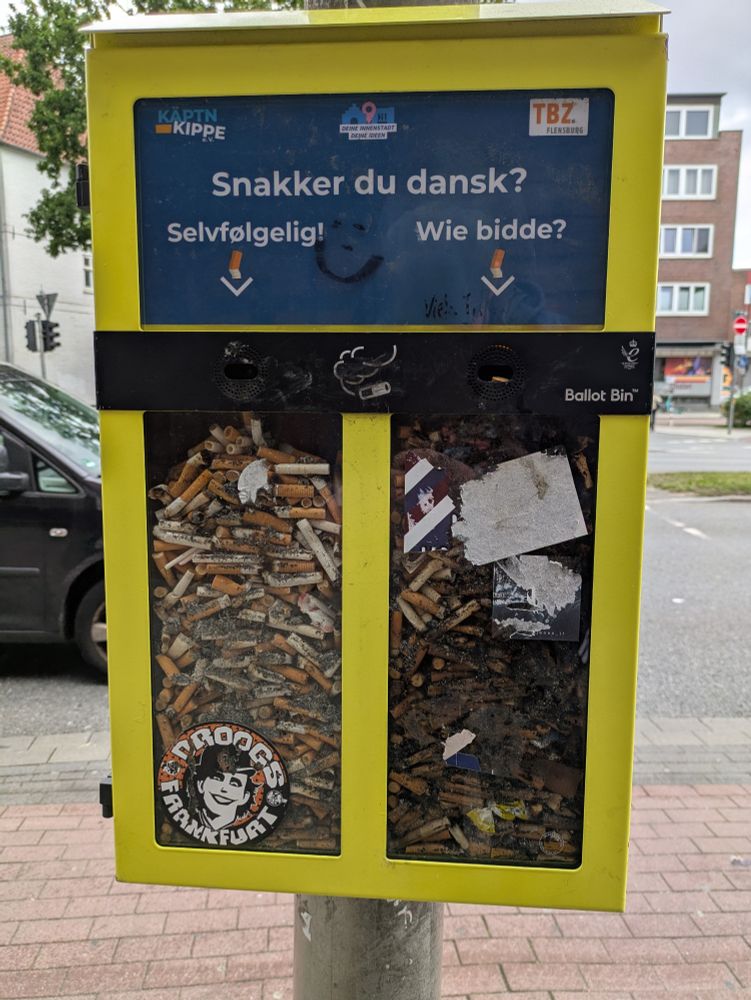 Gelber Kasten, angebracht an einem Ampelmast, mit zwei Schächten zum Einwerfen von Zigarettenkippen zum Votieren auf die Frage 