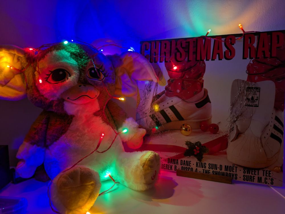 On the right side a record sleeve: "Christmas Rap" showing a pair of festively decorated feet in white Adidas sneakers. On the left: a plushie doll of Gizmo, the mogwai from "Gremlins". Both wrapped with a string of colourful fairy lights. 