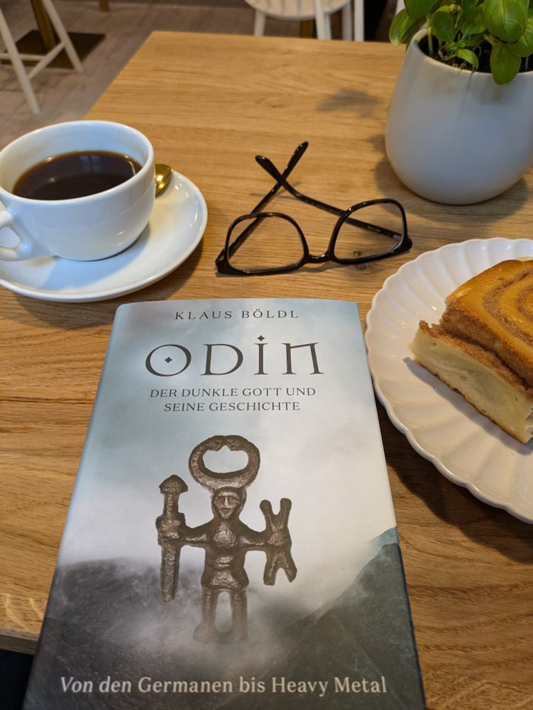 Tisch in einem Café, darauf arrangiert eine Tasse Kaffee, eine Brille und vor allem ein Buch: Klaus Böldl, "Odin - Der dunkle Gott und seine Geschichte. Von den Germanen bis Heavy Metal" (Verlag C. H. Beck). Das Titelbild zeigt eine Bronzestatuette (Odin?) mit gehörntem Helm und Schwert.