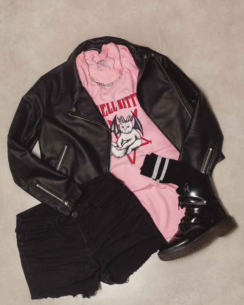 Look completo de roupas com jaqueta de couro camiseta rosa short preto meia preta com listras brancas e coturno sob um fundo acinzentado 
