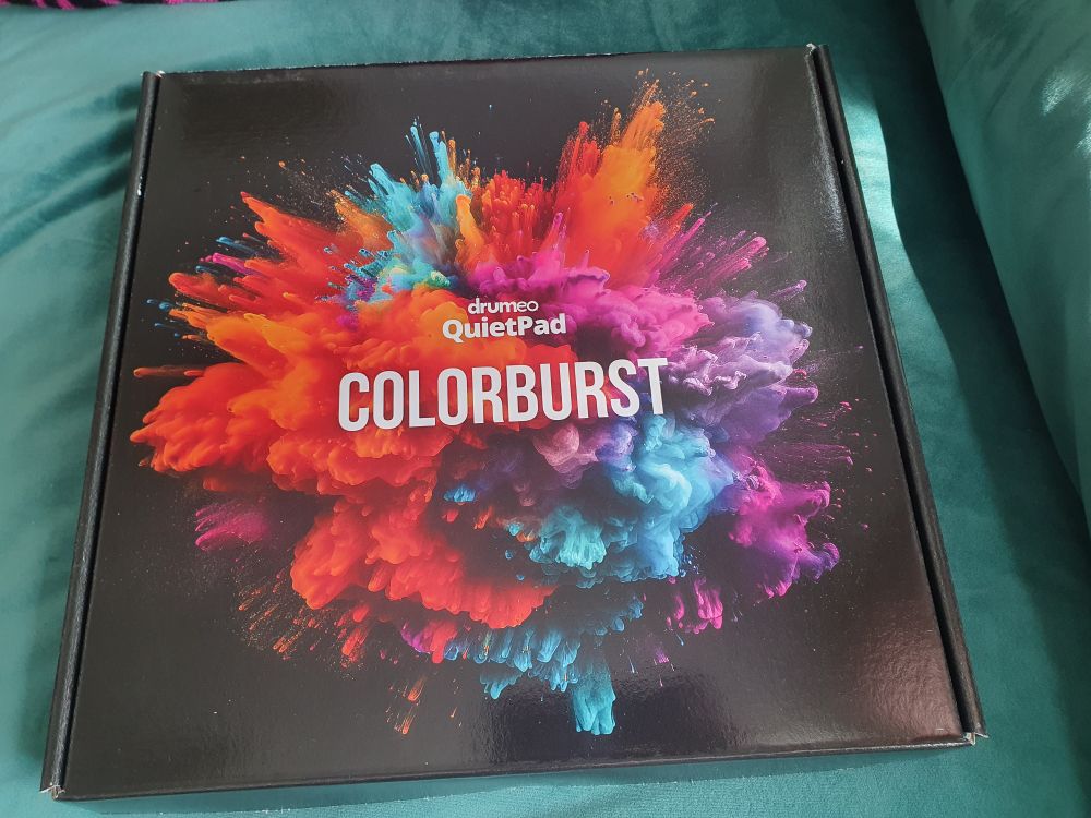 A box from Drumeo that contains a QuietPad. The box is black and has colorburst on the front with a stylised explosion of colour in orange, pink, blue and purple 