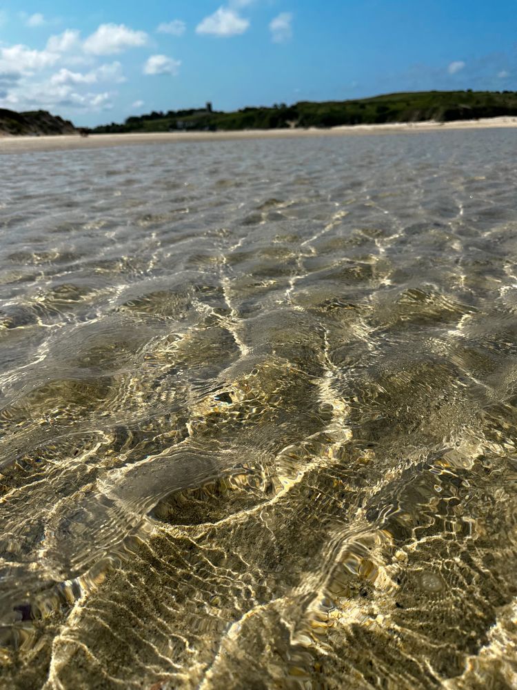 An image of the shallow sea near the shore, golden sand can be seen through the ripples in the water. On horizon green hills can be seen. 