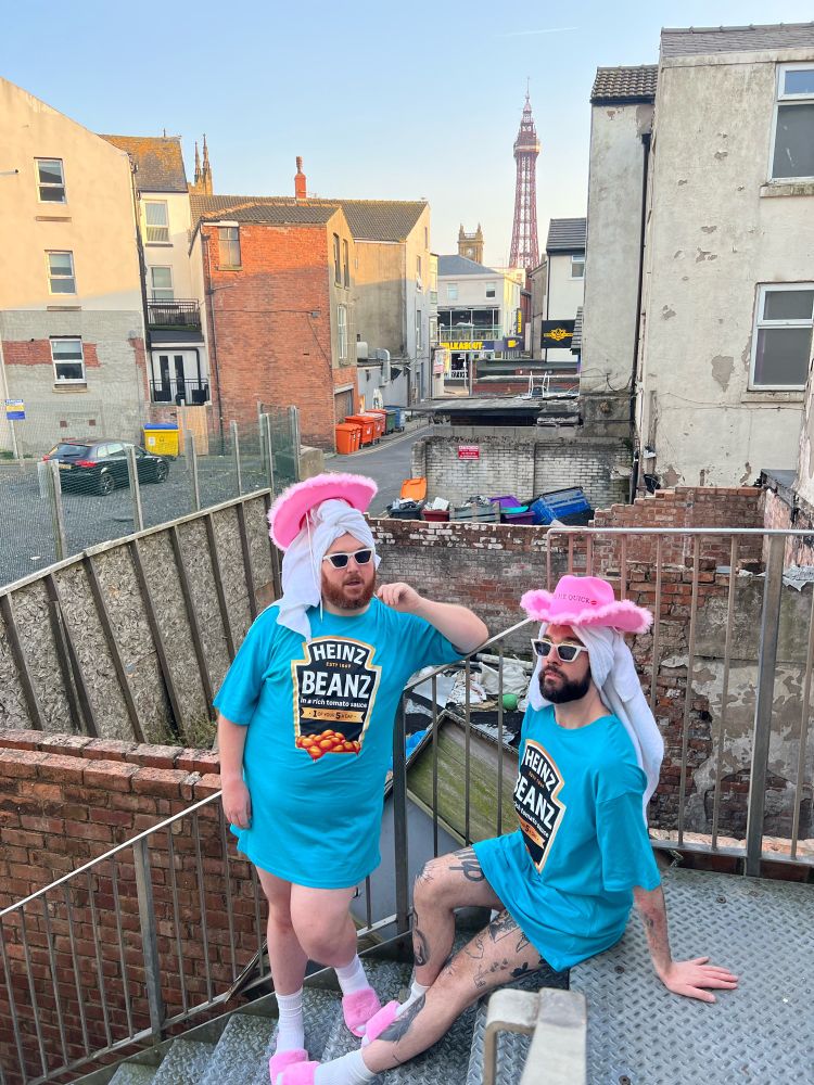 Harry and Scottee are wearing pink Kiss Me Quick cowboy hats, white towels wrapped around their head, Heinz Beanz nighties, white socks and pink furry slippers. They are on a metal fire escape with Blackpool Tower in the background. 