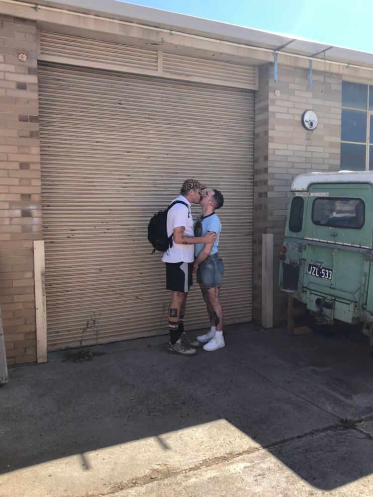 Two people kissing in front of a garage door by an old green car. One is wearing a leopard print cap, white shirt, backpack, black shorts and socks and walking boots. The other is wearing a blue t-shirt, denim shorts, white socks and shoes.  