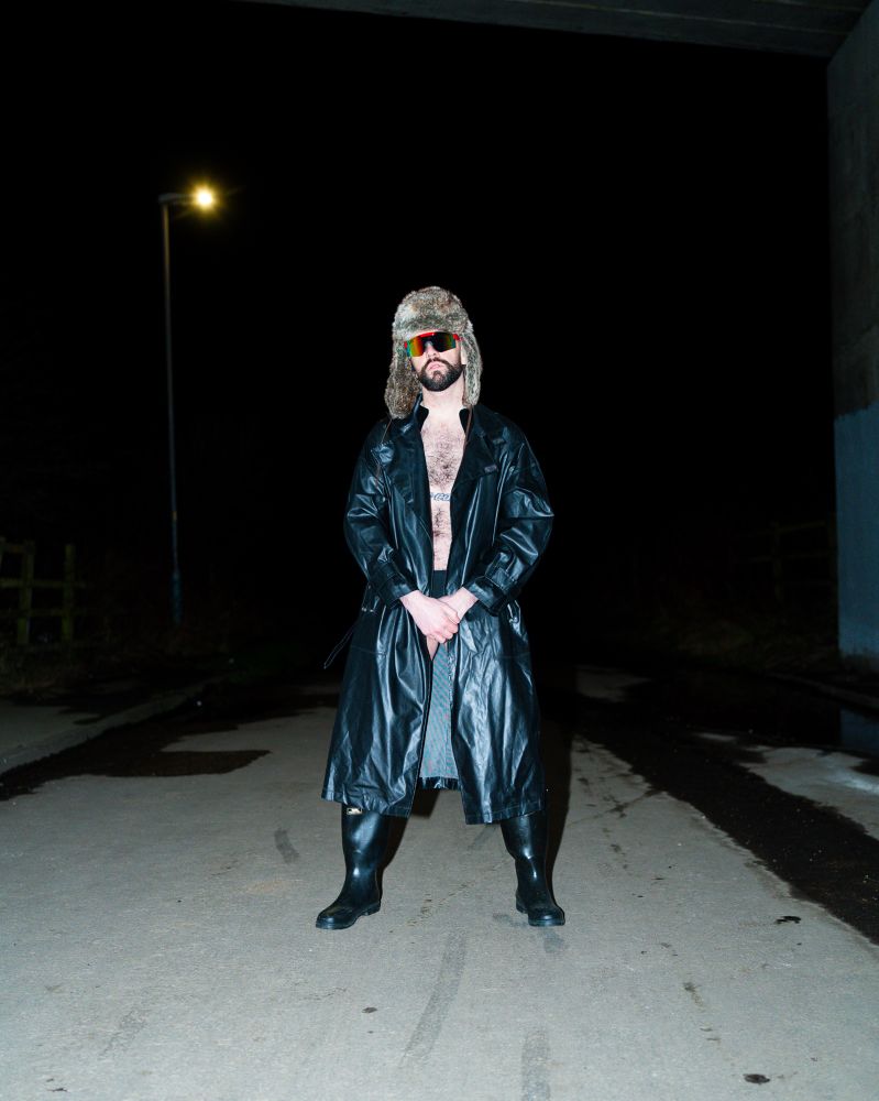 Harry in black trench coat, wellies, trapper hat and sunglasses. Black swim trunks peaking out as Harry holds fists in front of coat which is partially closed. High intensity flash. Harry under a bridge. 