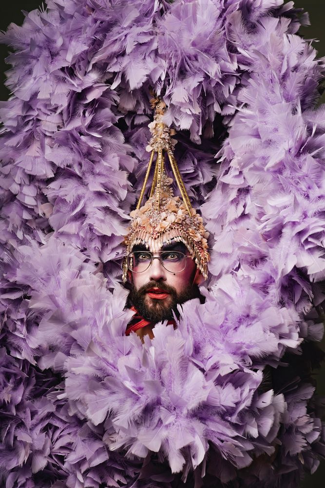 Mr Blackpool’s face, performed by Harry Clayton-Wright, is in the centre of a large clump of lilac feathers. They are wearing a giant headdress that resembles Blackpool Tower. A black beard, beauty mark, pale white powdered skin, dark eyeshadow and 70s aviator glasses.