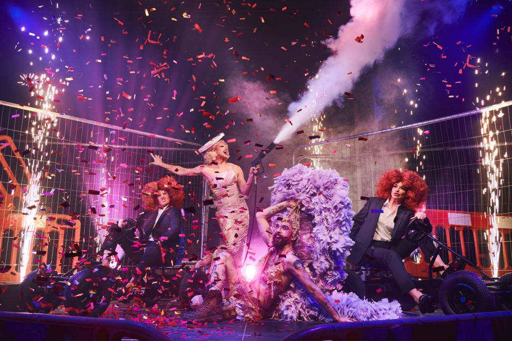 Four performers are on a stage in various positions. Two at either side of the image are dressed in drag as Mick Hucknall and are on go karts. One on the left of the image is pulling a face akin to a grimace and the other is giving fashion. They are in large wigs and black suits with white shirts. One performer at the back of the image in the centre left is stood up holding a CO2 gun and is dressed in drag as a stick of rock. The other performer at the front of the image centre right is laid on the floor in a giant lilac feather showgirl costume with one arm holding the headdress. Behind the performers is heras fencing, traffic panels and pyro. Throughout the image is confetti. The lights are lilac and it looks incredibly exciting. 