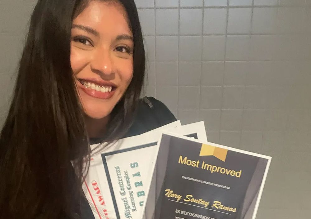 Nory Sontay Ramos holds a stack of school certificates, including one labeled “Most Improved,” and smiles for the camera.