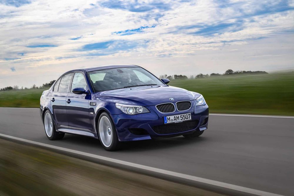 A press photo of a BMW e60 M5 in blue driving fast on a narrow country road with green grass and trees in the background. 