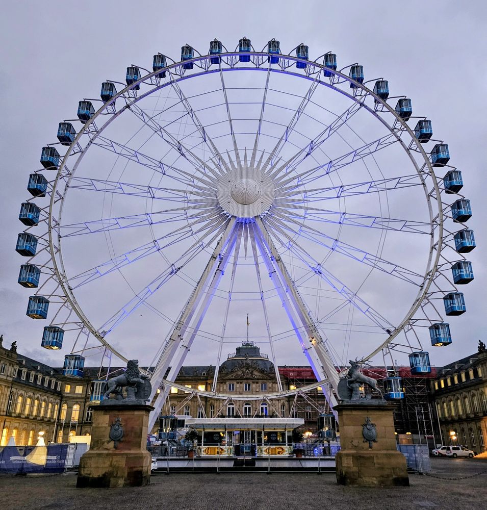 Auf dem Bild ist ein großes, hell beleuchtetes Riesenrad zu sehen, das zentral im Vordergrund steht und den Großteil des Bildes einnimmt. Die Gondeln sind geschlossen, überwiegend blau gefärbt und gleichmäßig entlang des weißen Stahlgerüsts angeordnet. Das Riesenrad ist mit dezenten blauen Lichtern illuminiert, die vor dem grauen, wolkenverhangenen Abendhimmel besonders hervorstechen.

Im Hintergrund befindet sich ein historisches, repräsentatives Gebäude mit klassischer Fassade und einer zentralen Kuppel, das von beiden Seiten von Flügelbauten eingerahmt wird. Es handelt sich um das neue Schloss in Stuttgart. Am unteren Bildrand stehen zwei steinerne Sockel mit Skulpturen, links ein Löwe, rechts ein Hirsch, die den Eingang zum Riesenrad flankieren. Um die Anlage herum sind Absperrgitter, Bauzäune und Beleuchtungselemente zu erkennen. 