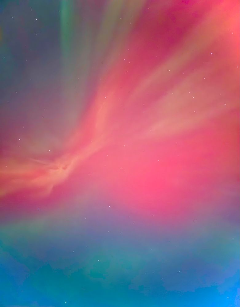 A view of the northern lights from directly below. A few stars peak through the pinks and greens that fork out from the center like a watercolor painting of a synapse