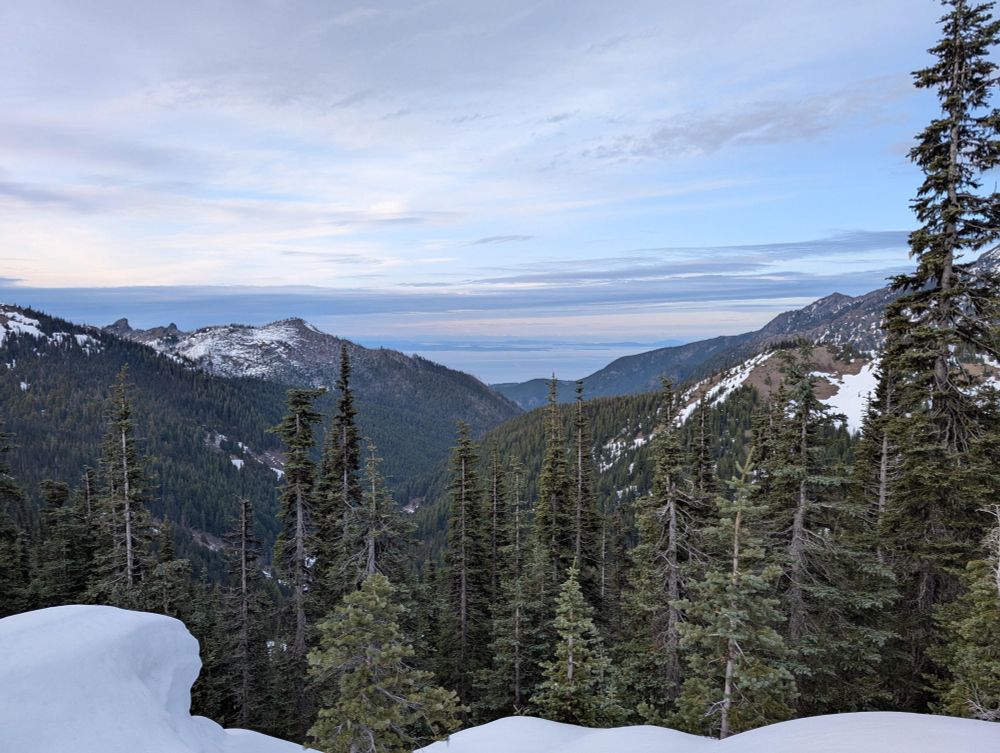 Photo from high elevation of snowy mountain slopes framing an evergreen rain forest valley leading north down to the Salish Sea, with Vancouver Island and the city of Victoria distantly in the haze.