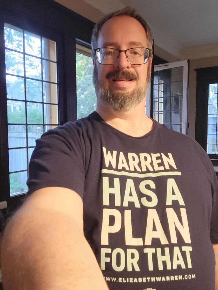 Me, a 50-year-old bearded white dude with glasses, wearing a dark blue tee shirt that says "Warren has a plan for that" in capital white and light teal letters.