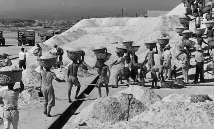 B&w still of salt hills with workers walking around with baskets over their heads.