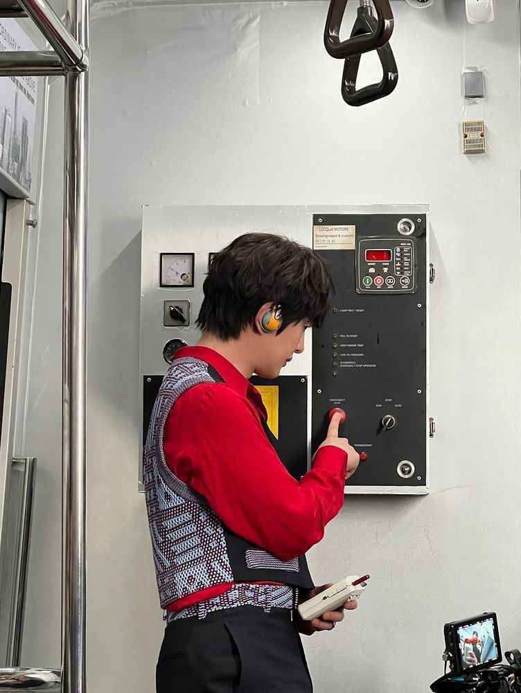Onew pressing a button on the metro car control panel. He’s holding a small device in his hand and wearing headphones 