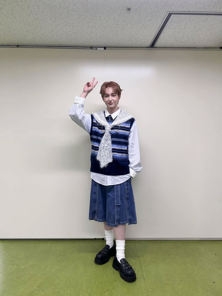 Jinki wearing a soft striped sweater vest over denim shorts, holding his right hand above his head in a v-sign