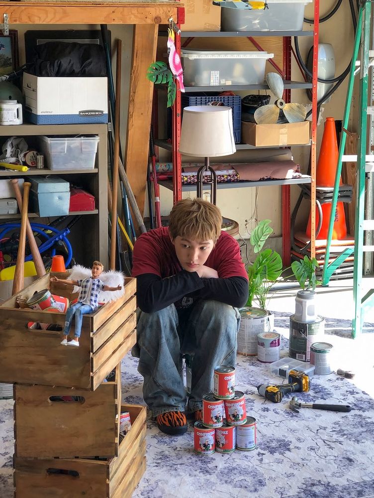 Onew sitting on the floor next to the spinach crates, knees drawn up and chin resting on his crossed arms 