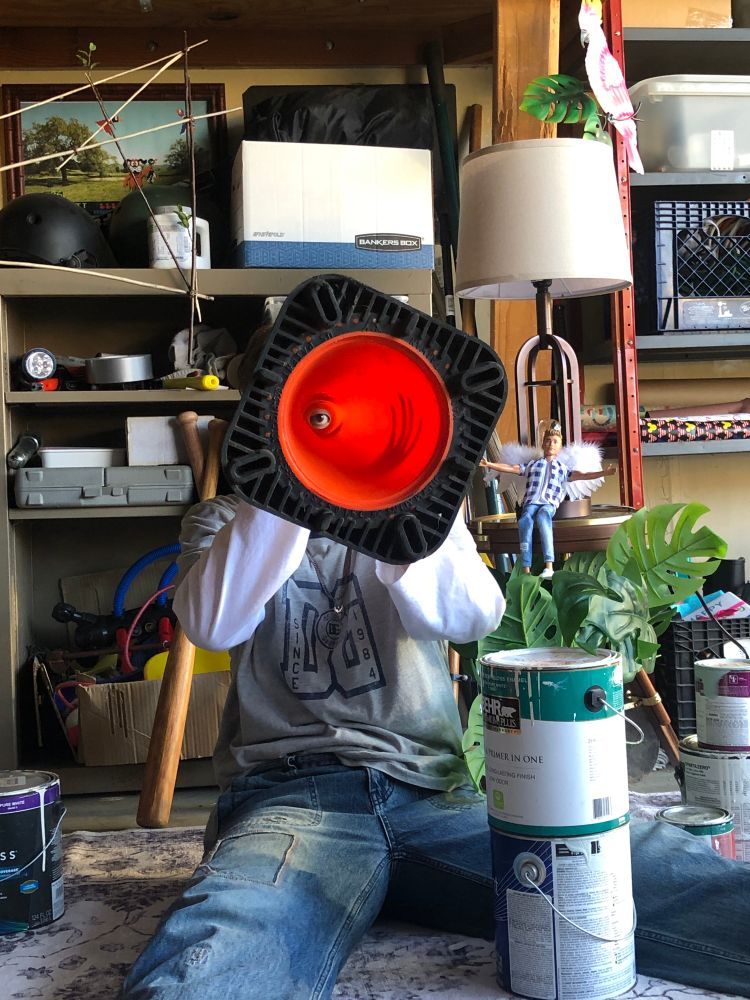 Onew sitting on the floor and holding an orange construction cone in front of his face. He’s surrounded by shelves and home maintenance items like paint and tools. There’s also a Ken doll with angel wings sitting near him 