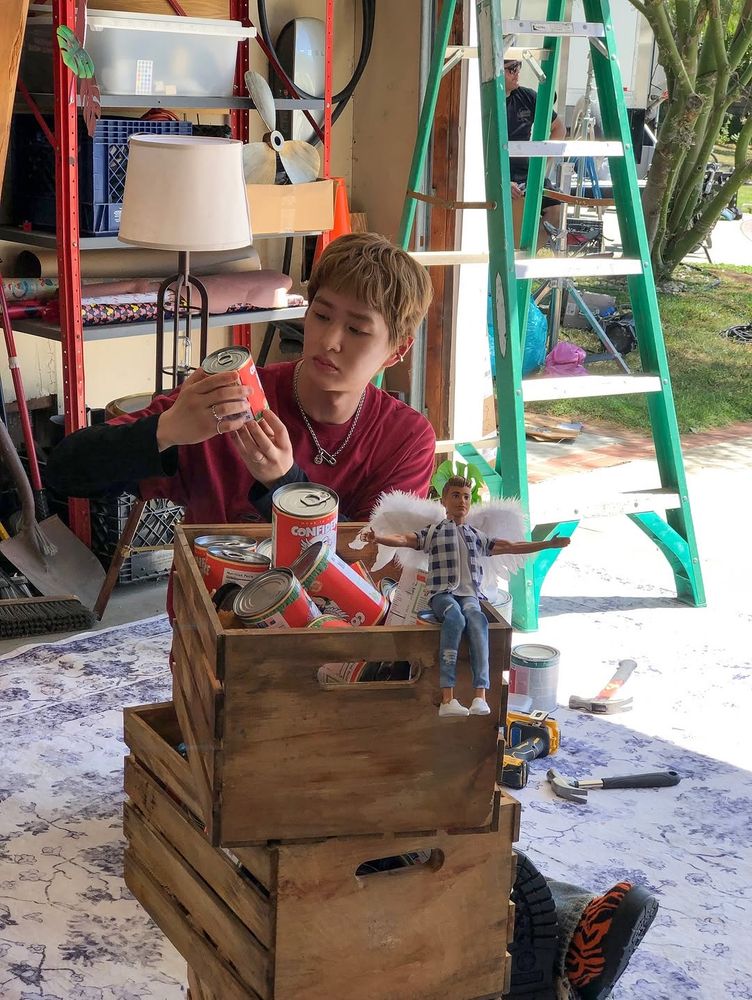 Onew sitting on a garage floor with three wooden crates stacked in front of him. There are cans of spinach in the crates and a Ken doll with wings sitting on the edge of the top crate. Onew is wearing jeans and a red tshirt over a black longsleeved tshirt and inspecting a can of spinach that he’s holding in his hand 
