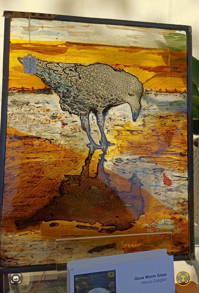 A raven is painted onto glass in hues of black, grey, blue and golden orangey yellow. Its reflection is clear and looks to be on wet sand.