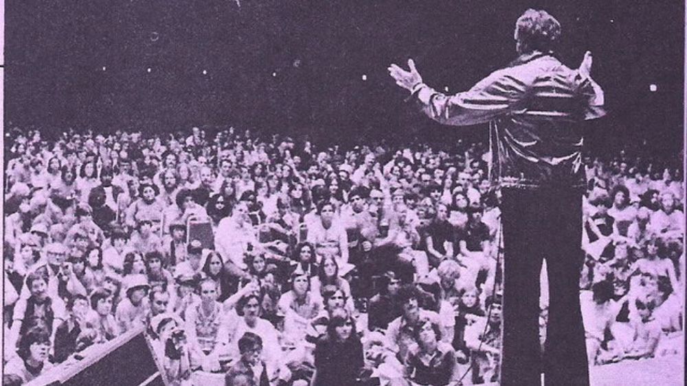 Newspaper photo of a large crowd watching a ma on the stage at an early Star Trek convention, possibly even the first in 1976 at the Statler-Hilton Hotel in NY. Even though the resolution is not great you can still easily make out many female-presenting individuals. 