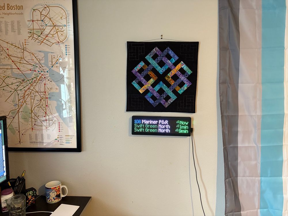 The wall of a home office, showing an electronic sign board listing three bus arrivals.  There’s also a poster with a transit map of Boston, a Celtic knot embroidery, and a demiboy pride flag visible.