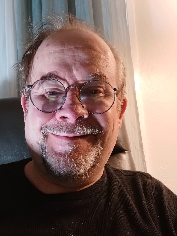 A picture of Jeff Strasburg, a man with a grey beard and sandy brown hair wearing glasses, and a black t-shirt. He's sitting on a charcoal grey recliner. There are green and off-white curtains hanging behind him and to his left, and a bare light grey wall to his right.