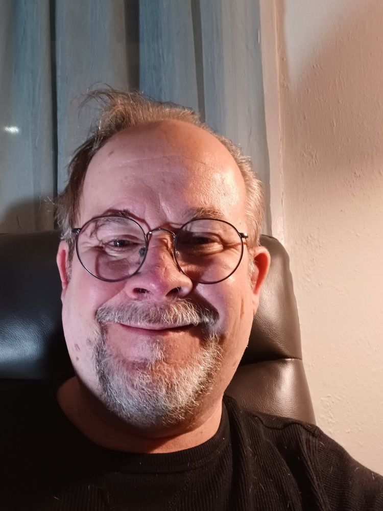 A picture of Jeff Strasburg, a man with a grey beard and sandy brown hair wearing glasses, and a black t-shirt. He's sitting on a charcoal grey recliner. There are green and off-white curtains hanging behind him, and a light grey wall to his right.
