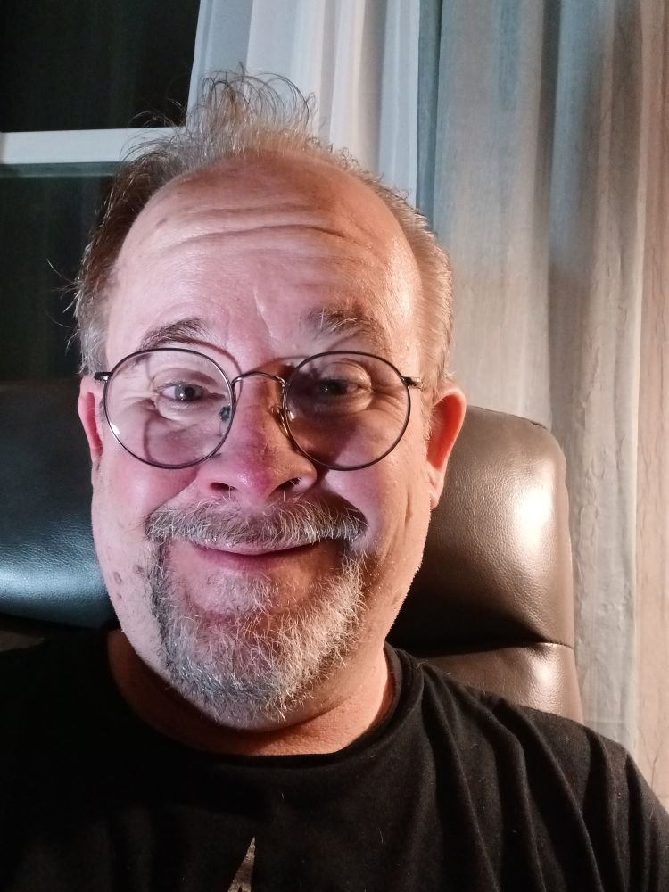 A picture of Jeff Strasburg, a man with a grey beard and sandy brown hair wearing glasses, and a black t-shirt with a screen-printed silver fleur de lis. He's sitting on a charcoal grey recliner. There are green and off-white curtains hanging behind him and to his right, and a dark window behind him to his left.