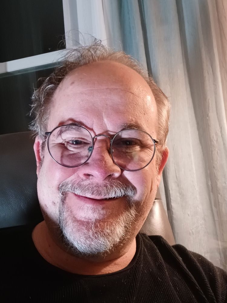 A picture of Jeff Strasburg, a man with a grey beard and sandy brown hair wearing glasses, and a black t-shirt. He's sitting on a charcoal grey recliner. There are green and off-white curtains hanging behind him and to his right, and a dark window to his left.