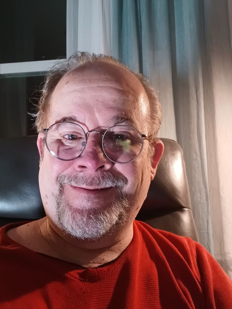A picture of Jeff Strasburg, a man with a grey beard and sandy brown hair wearing glasses, and a red v-neck t-shirt. He's sitting on a charcoal grey recliner. There are green and off-white curtains hanging behind him and to his right, and a dark window to his left.