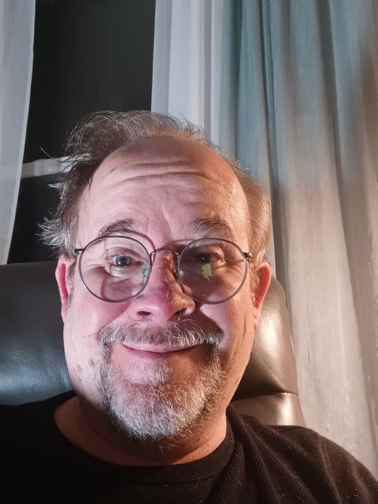 A picture of Jeff Strasburg, a man with a grey beard and sandy brown hair wearing glasses, and a black t-shirt. He's sitting on a charcoal grey recliner. There are green and off-white curtains hanging behind him and to his right, a white curtain to his far left, a dark window in between them.
