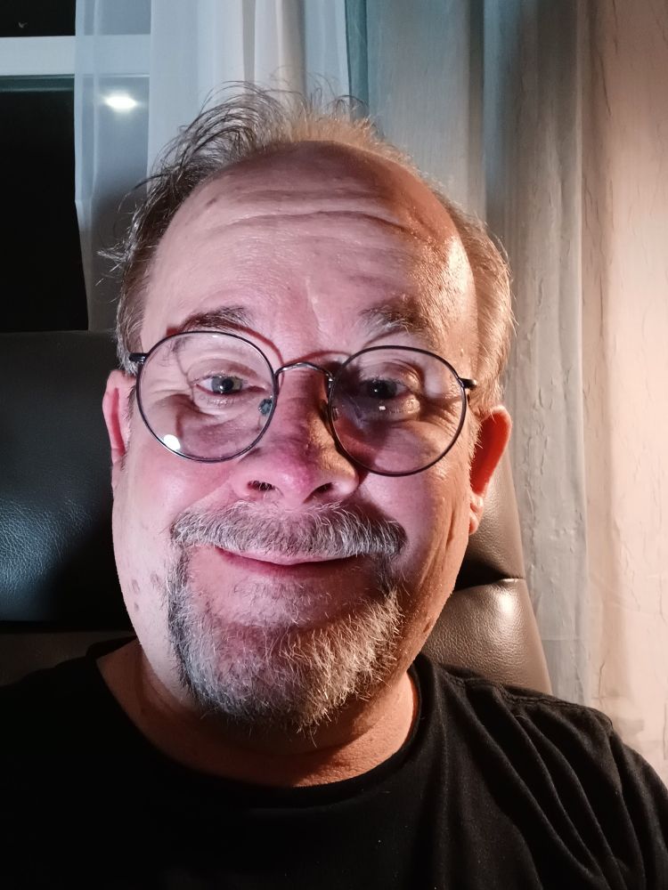 A picture of Jeff Strasburg, a man with a grey beard and sandy brown hair wearing glasses, and a black t-shirt with a screen-printed silver fleur de lis. He's sitting on a charcoal grey recliner. There are green and off-white curtains hanging behind him and to his right.