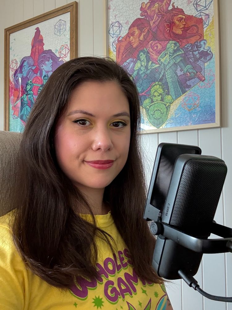 Bilde av Tanya Pettersen. Hun har på seg en gul t-skjorte fra Wholesome Games og sitter foran en sort mikrofon. Det henger fargerike, fine kunstverk basert på spill på veggen bak henne.