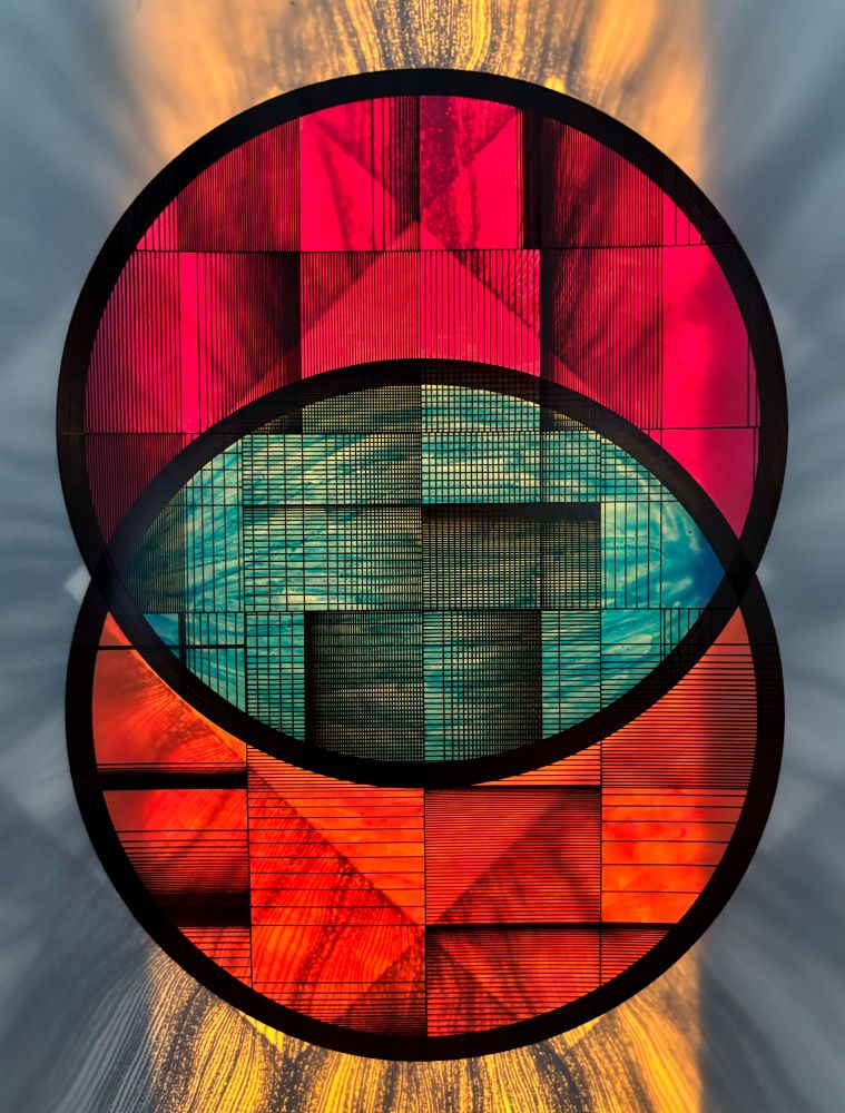 Two overlapping circles filled with intricate black generative linework appear over a vibrant, backlit surface. The top circle glows red and magenta, the bottom radiates fiery orange, while the overlapping center reveals cooler teal hues. Created on translucent cel with plotted ink and painted reverse, the piece evokes a stained-glass-like effect.