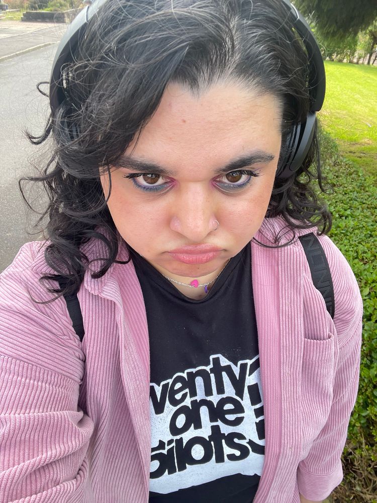 a selfie of me at a high angle, i'm frowning and wearing a black and white twenty one pilots shirt with a pink jacket and headphones, with blue, purple and pink eye makeup