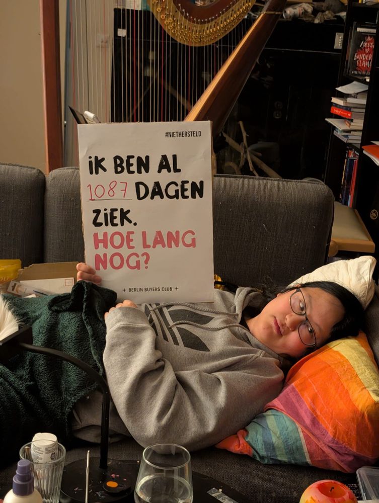 A picture of Fen lying on the couch. Their head is supported with a bed pillow. They are holding a sign that says 'ive been ill for 1087 days, how much longer?'