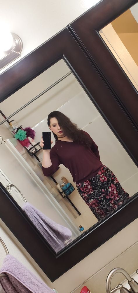 Ashley, post transition. She's wearing a black skirt with red and pink floral patterns strewn across the article. She has on a maroon top that floats lazily. Her hair is notably long and pretty.