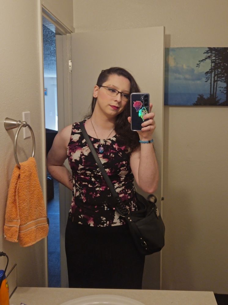 Ashley taking a mirror selfie. dressed up all pretty with a floural black, white and magenta top and long black skirt. She has on glasses and a hair tie sits around her wrist.